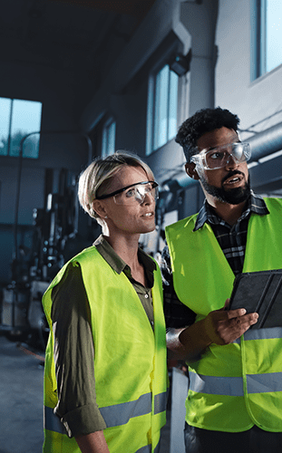 Two workers in a factory wearing high-visibility vests and safety glasses, examining a tablet together.