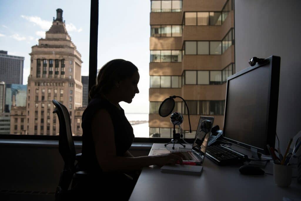 A silhouette of a woman seated at a desk working on a computer with a city buildings viewable from a high office window.