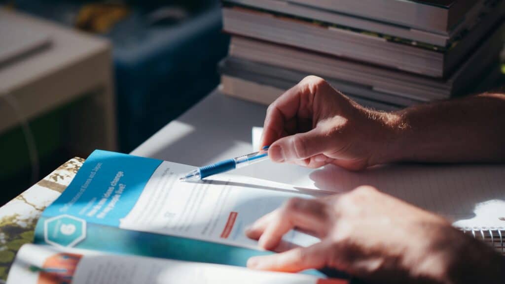 A person's hands holding a pen and pointing at a page from a thick manual-like textbook.