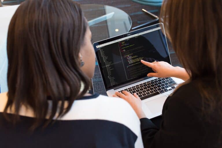 Two businesswomen look at code together on a laptop