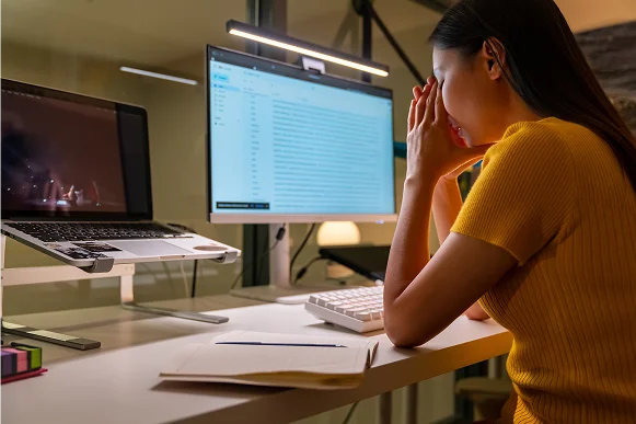 A woman in a yellow shirt sits at a desk, head in hands, overwhelmed by work on dual computer screens and papers nearby.