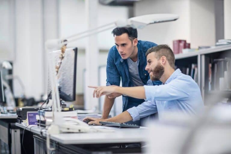 Two employees collaborating on a computer with IT support, discussing technology solutions in a modern office setting.