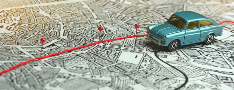 Blue toy car on a maps with red string tracing a route and pink pins marking stops.