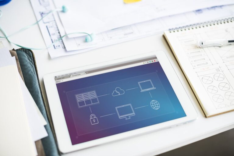 Tablet on a white desk displaying blueprint-style icons, with sketches, a notebook, and office supplies around it