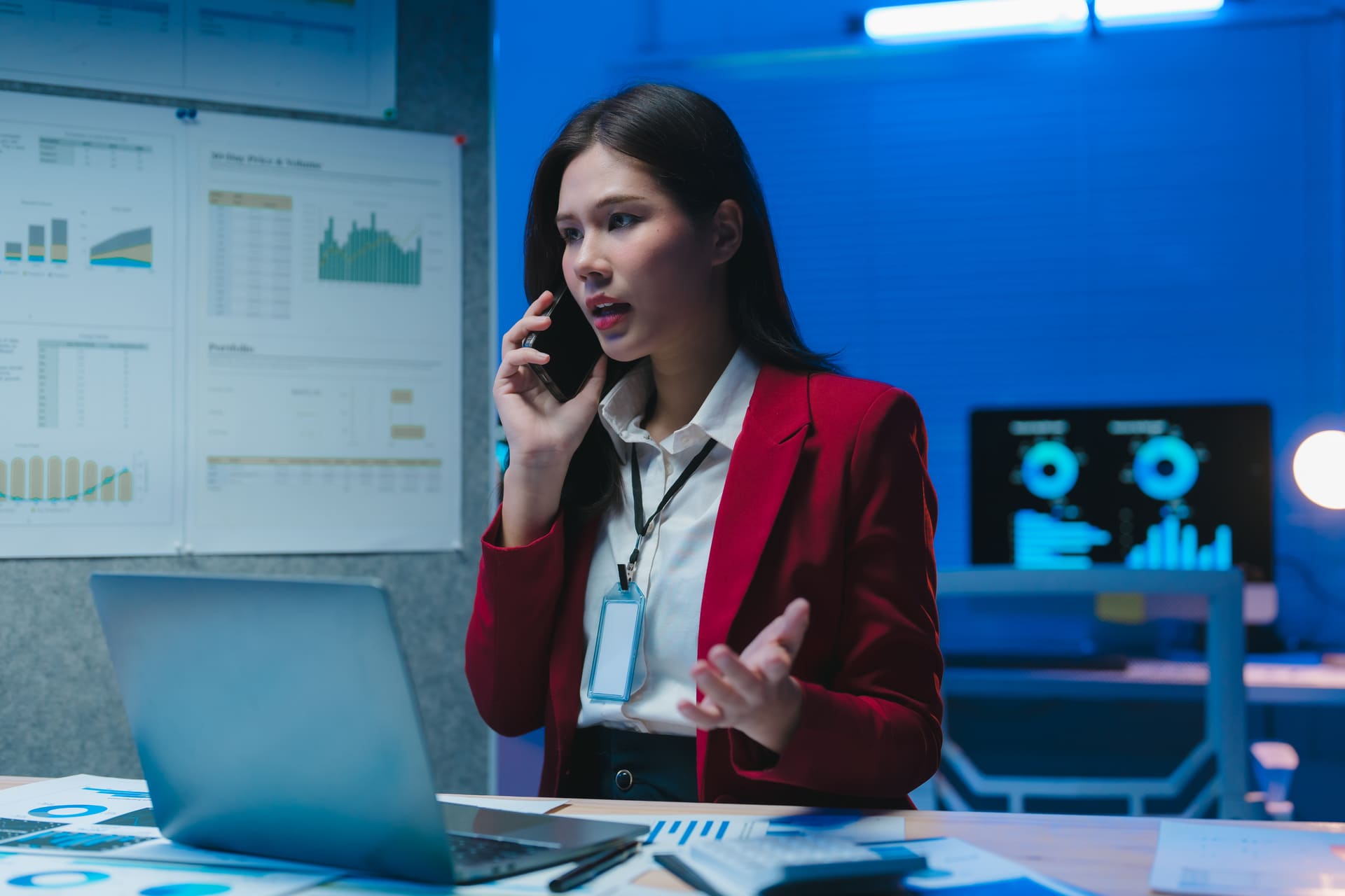 Businesswoman in a red jacket on the phone while working on a laptop with charts in the background