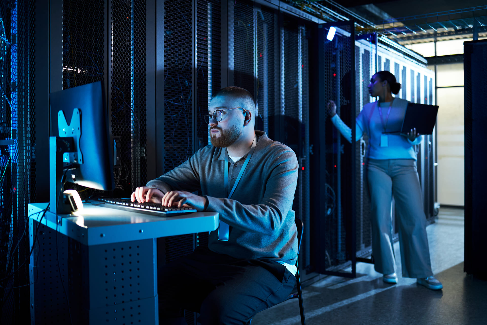 IT technician working on a computer in a server room while a colleague checks equipment