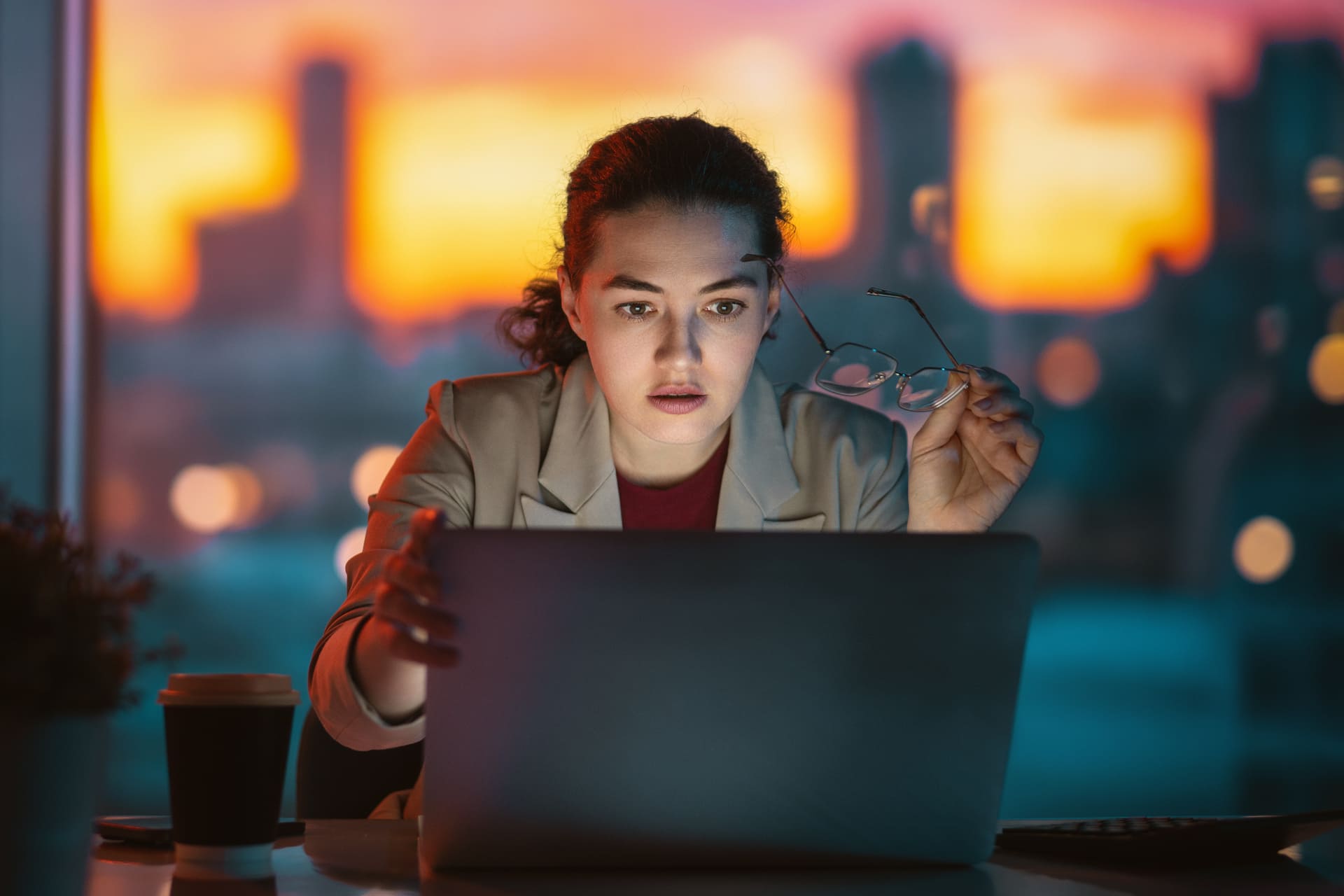 Woman working late on laptop with cityscape at sunset