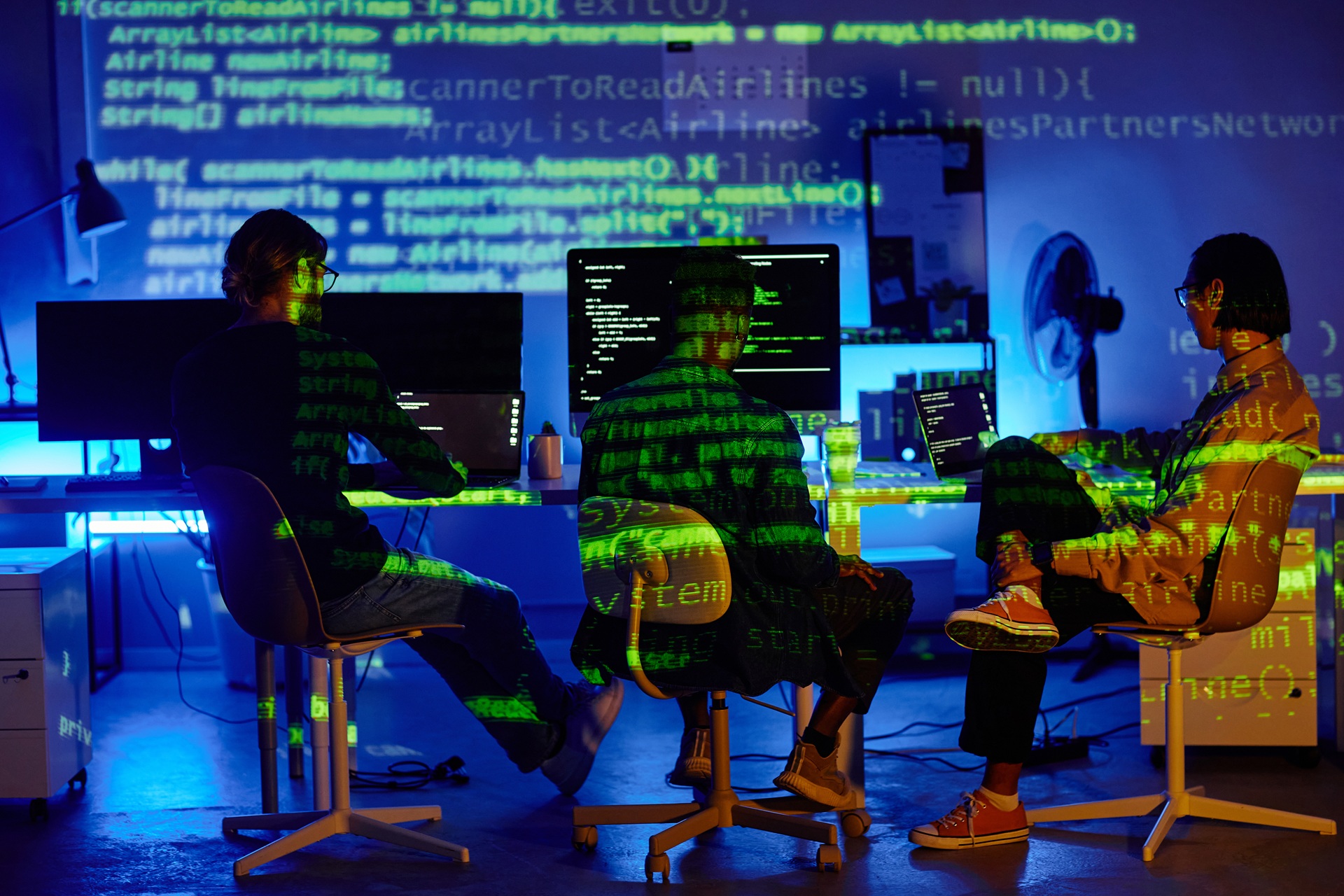 Group of programmers working in a dark room with green code projected onto their bodies and walls, surrounded by multiple monitors