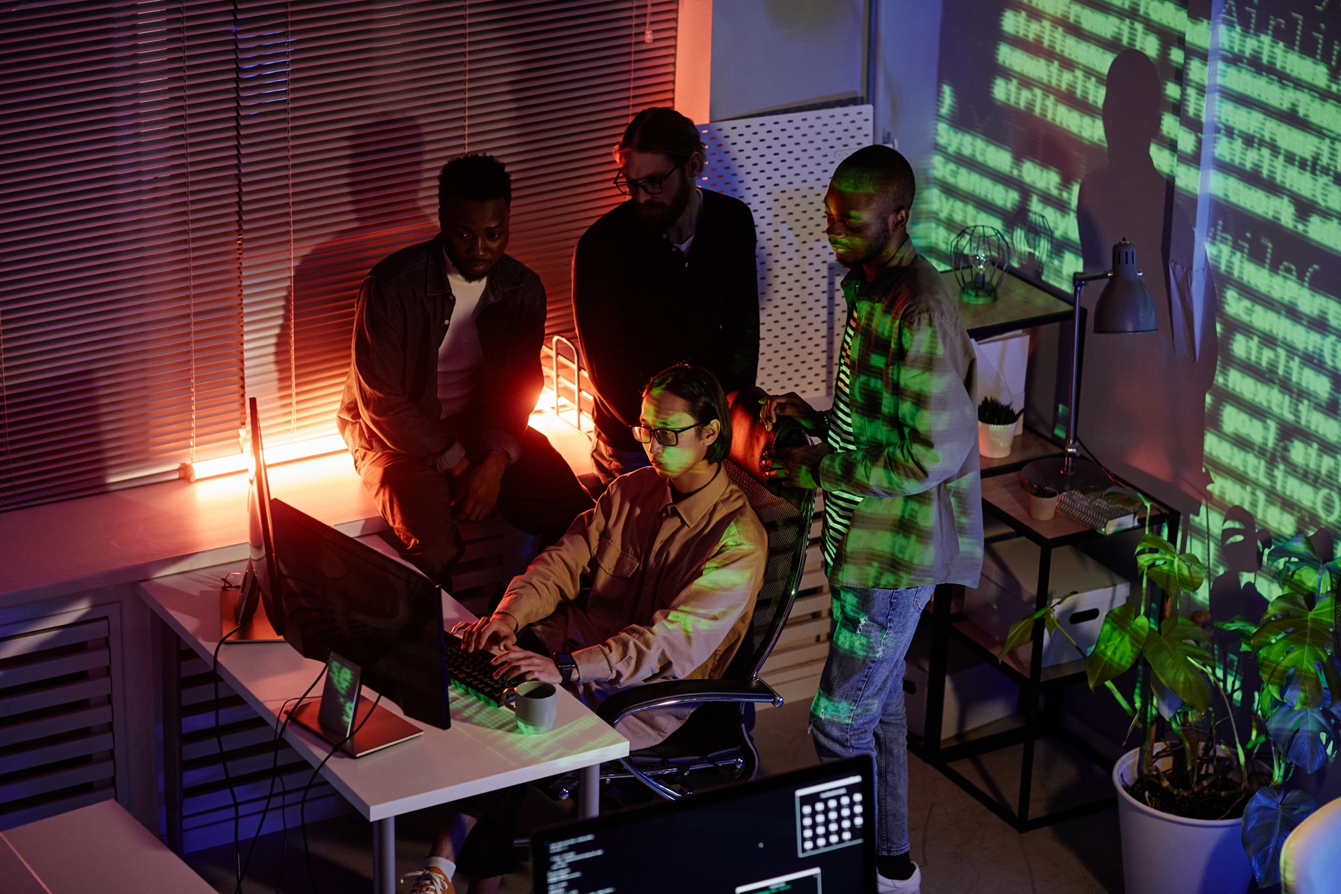 Group of young software developers gathered around a workstation, illuminated by code projections, working on cybersecurity or programming tasks