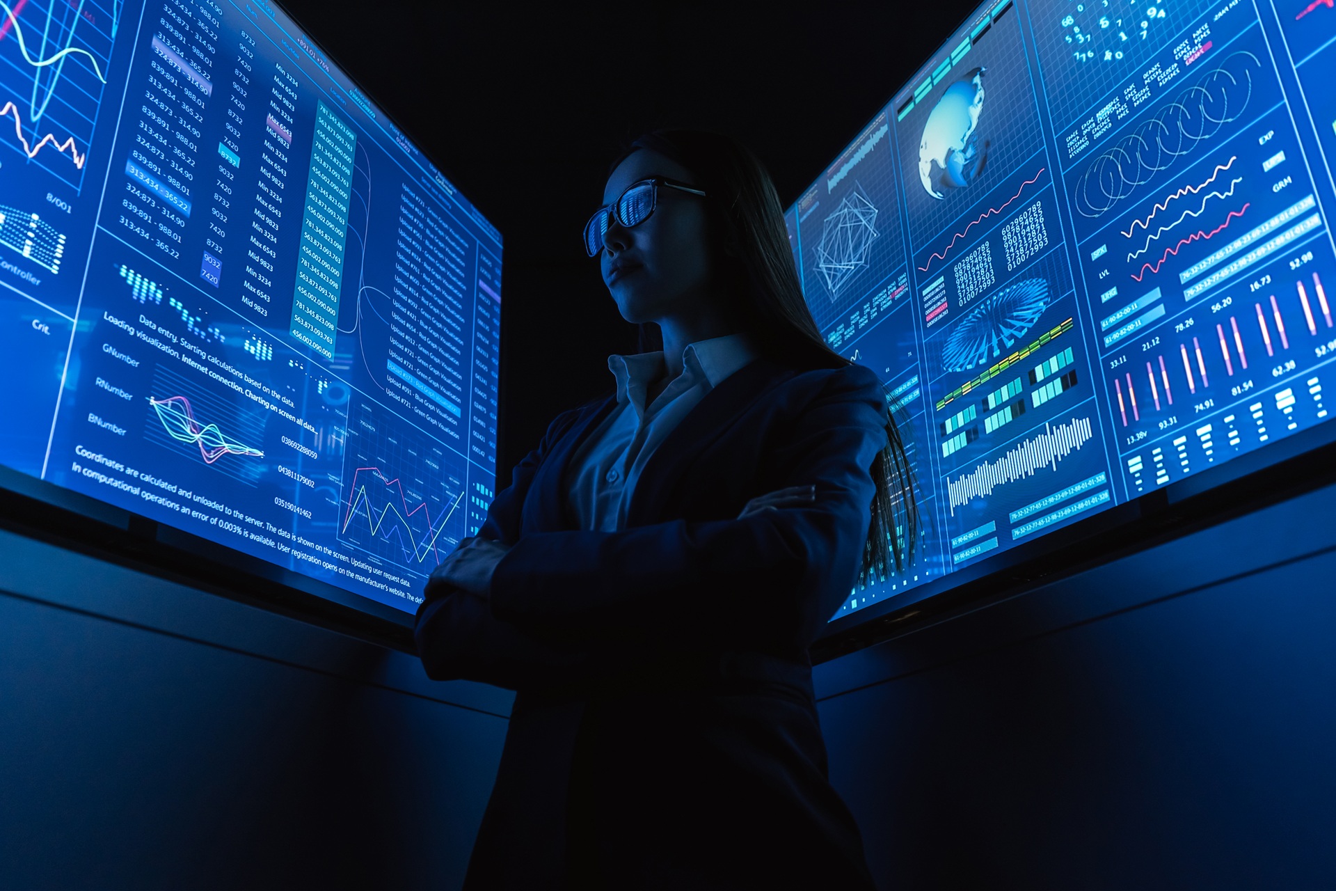 Businesswoman standing confidently with arms crossed in front of multiple large digital screens displaying data charts and analytics.