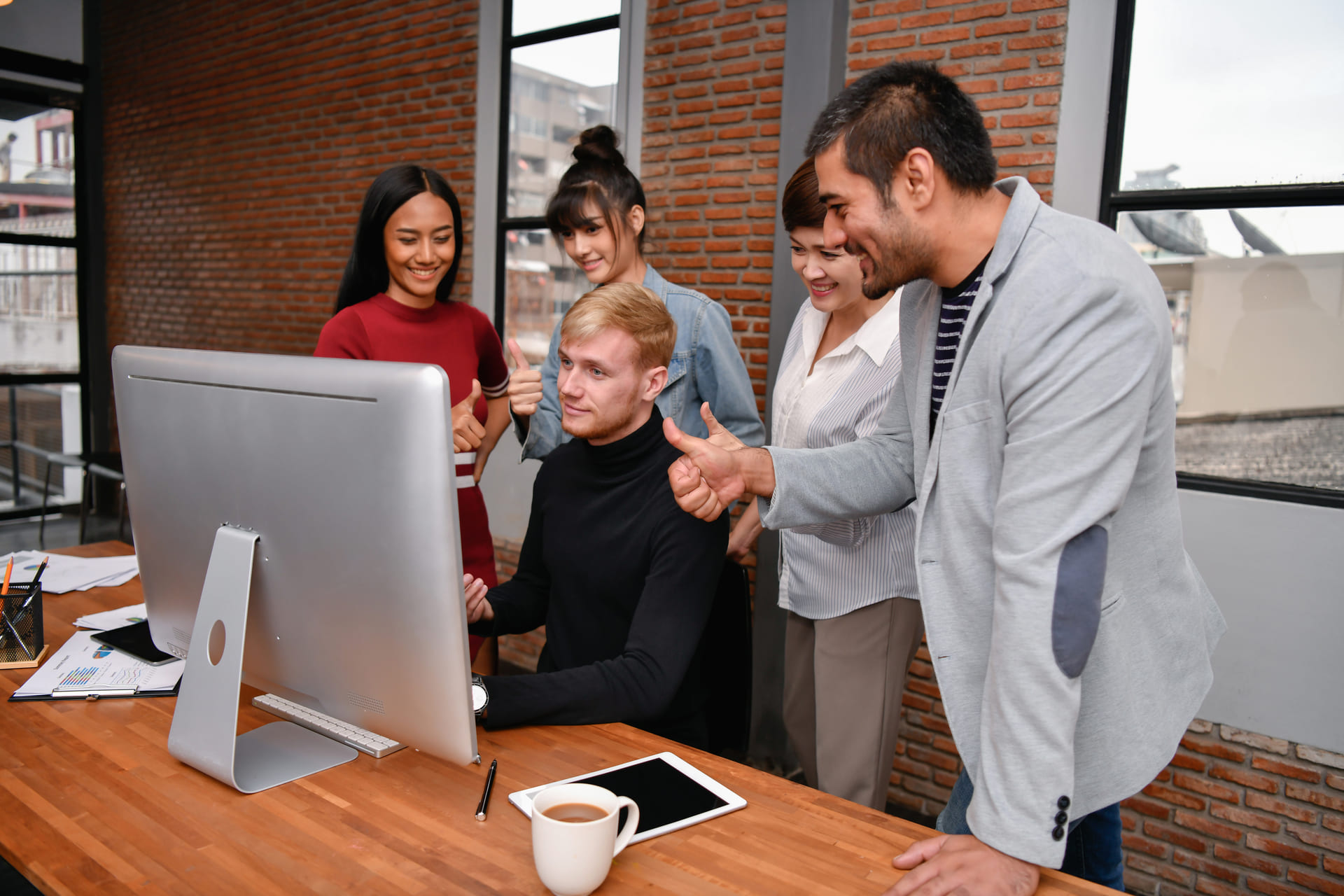 Group of coworkers giving thumbs up to a colleague at computer in modern office