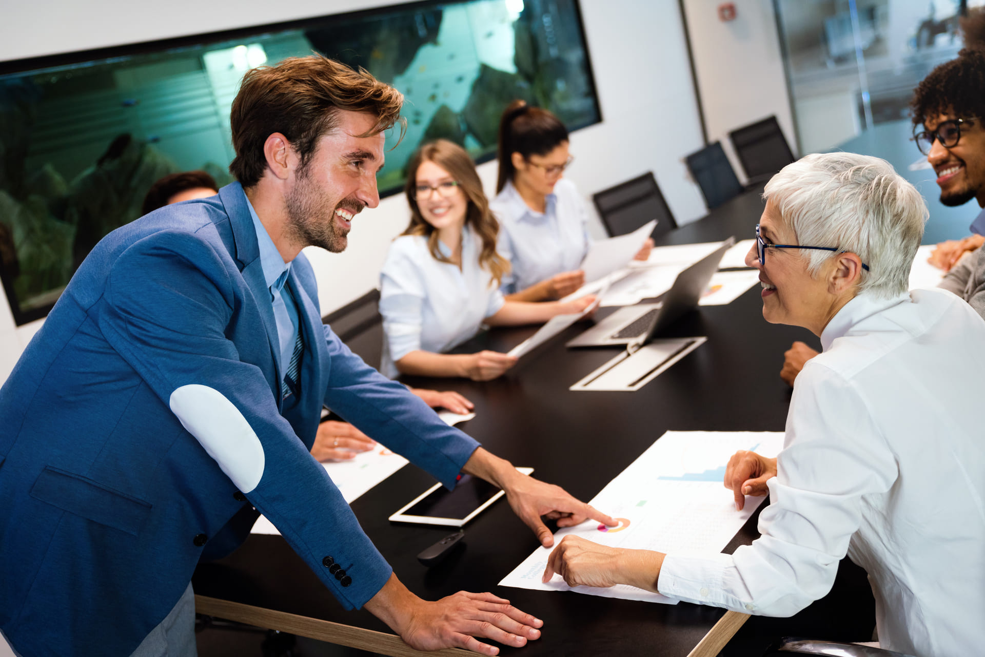 Diverse team in a meeting smiling and reviewing printed charts around a conference table | CMIT Solutions Diverse team in a meeting smiling and reviewing printed charts around a conference table