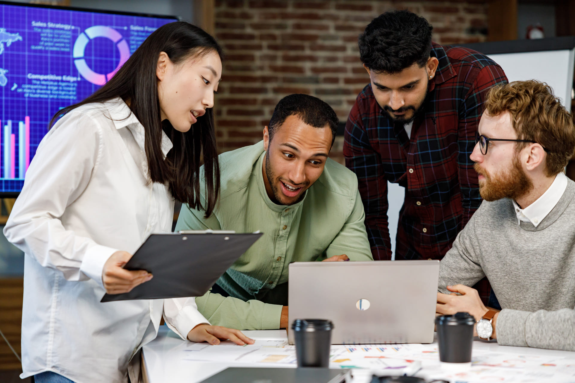 Four coworkers gathered around a laptop discussing charts and documents in an office | CMIT Solutions Four coworkers gathered around a laptop discussing charts and documents in an office