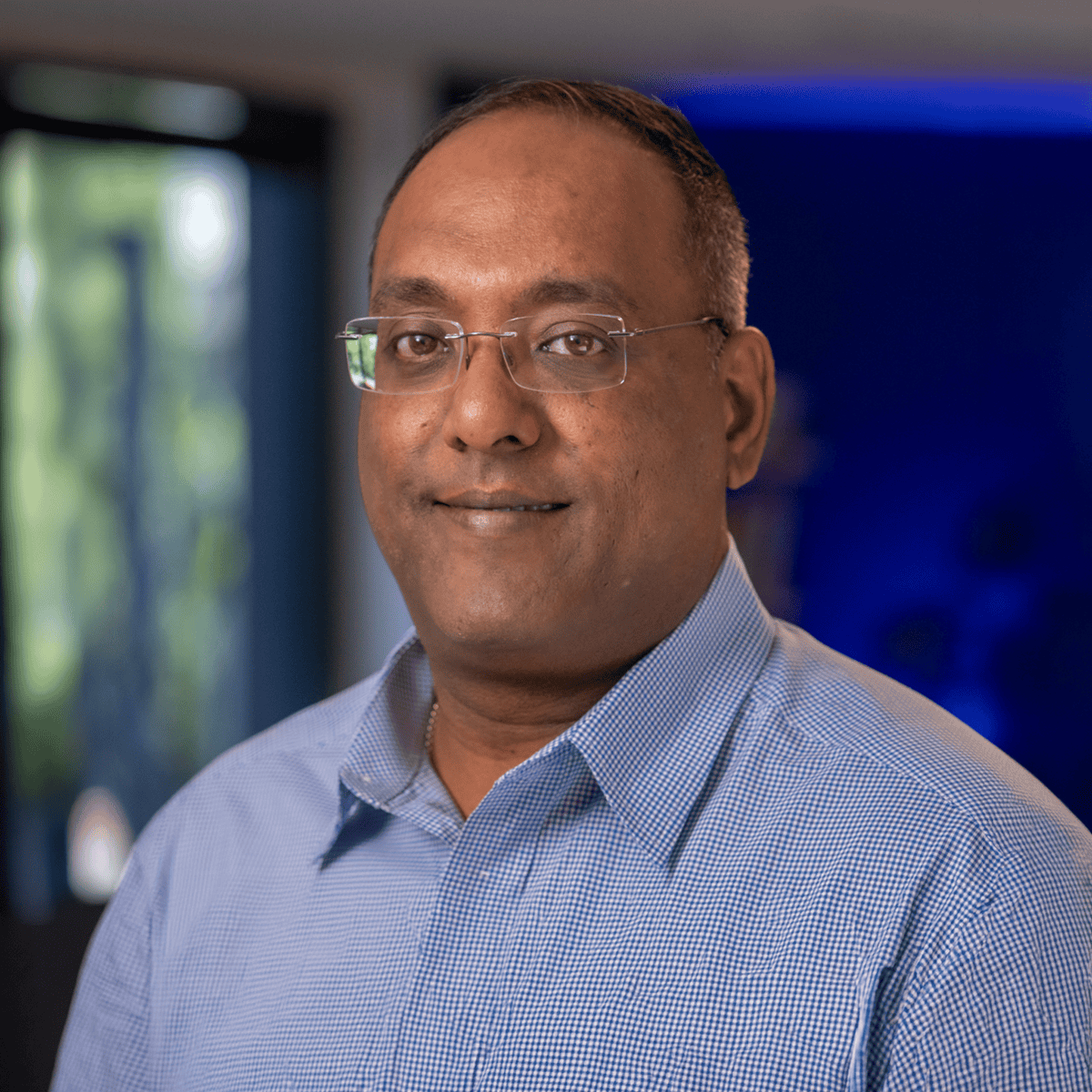Portrait of a man wearing glasses and a light blue checkered shirt, looking at the camera in an indoor setting.