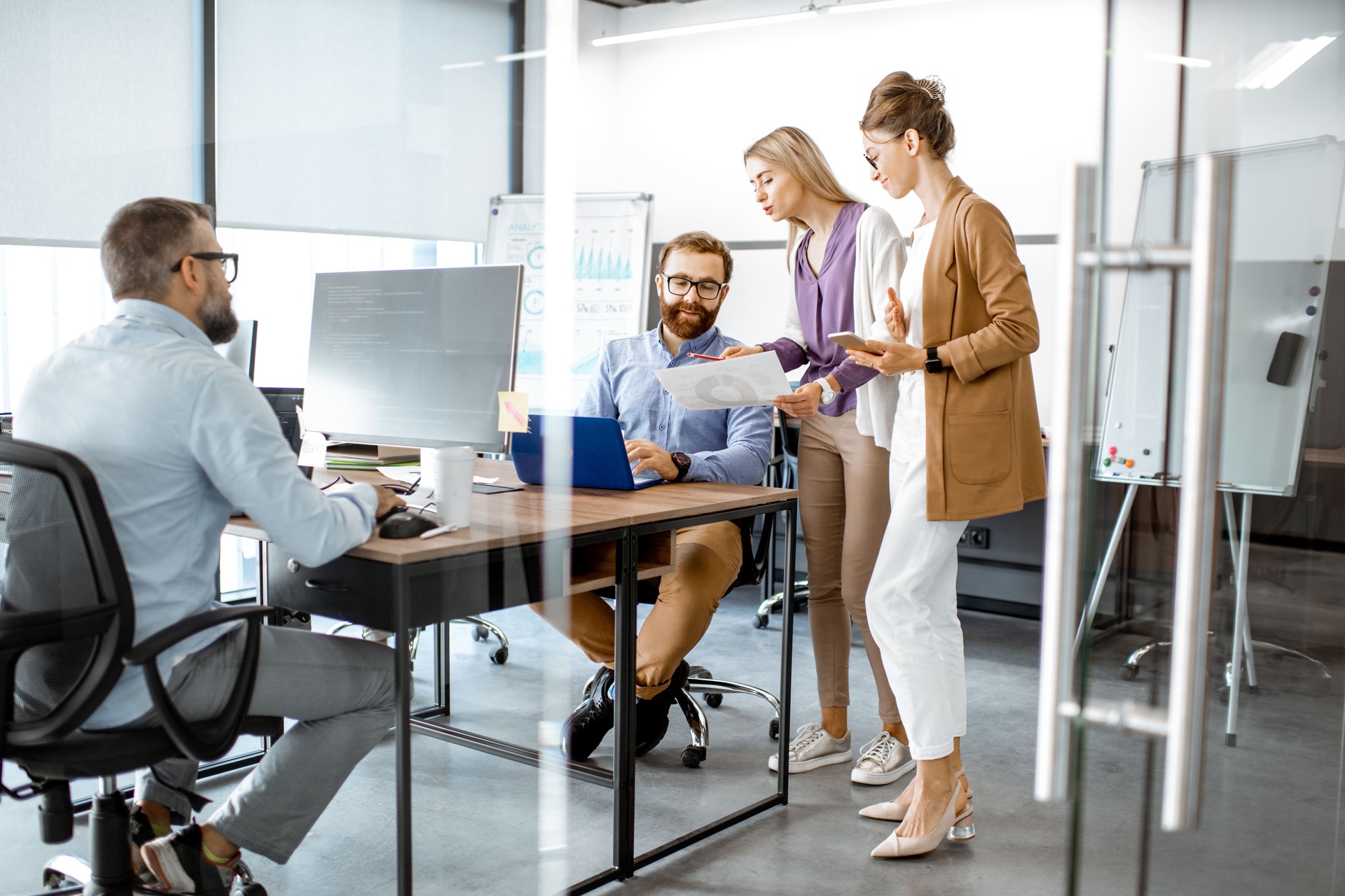 Programmers working at the office | CMIT Solutions Four colleagues in a modern office review a document together at a shared desk, one with a laptop open nearby.
