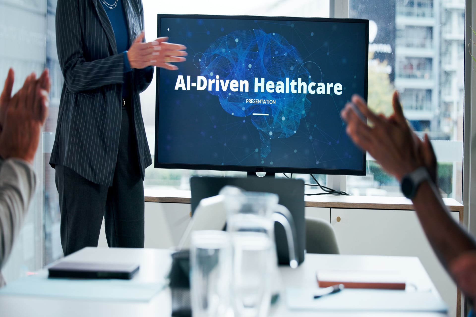 ai driven healthcare presentation on screen | CMIT Solutions Presenter standing beside a monitor that reads'AI-Driven Healthcare' during a presentation, audience applauding in a boardroom.