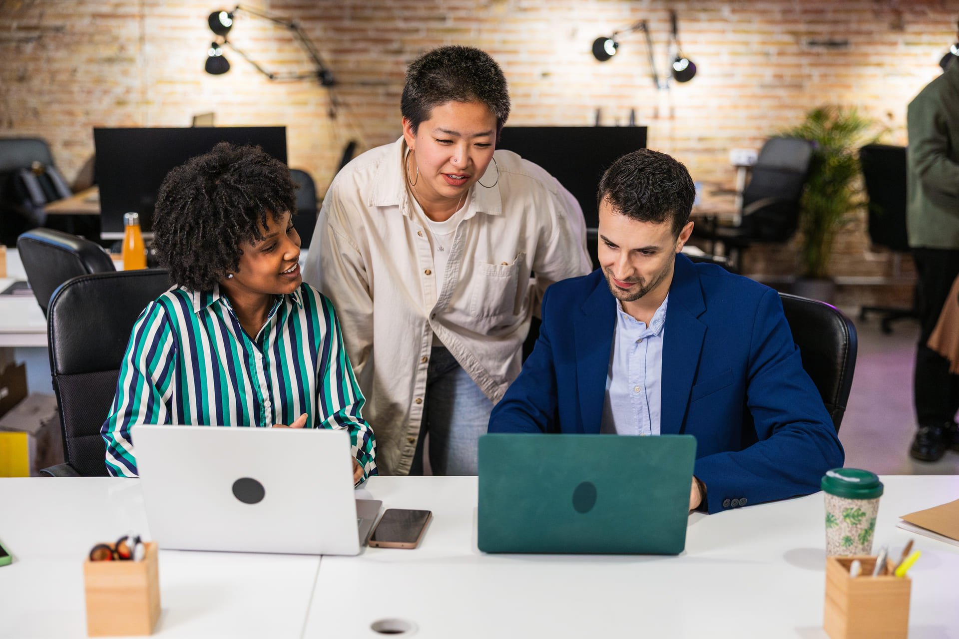 multi-ethnic-team-collaborating-on-project-using-laptops | CMIT Solutions Three colleagues collaborate at a white desk, two seated with laptops while a third stands, looking at the screen in a bright office with a brick wall behind them.