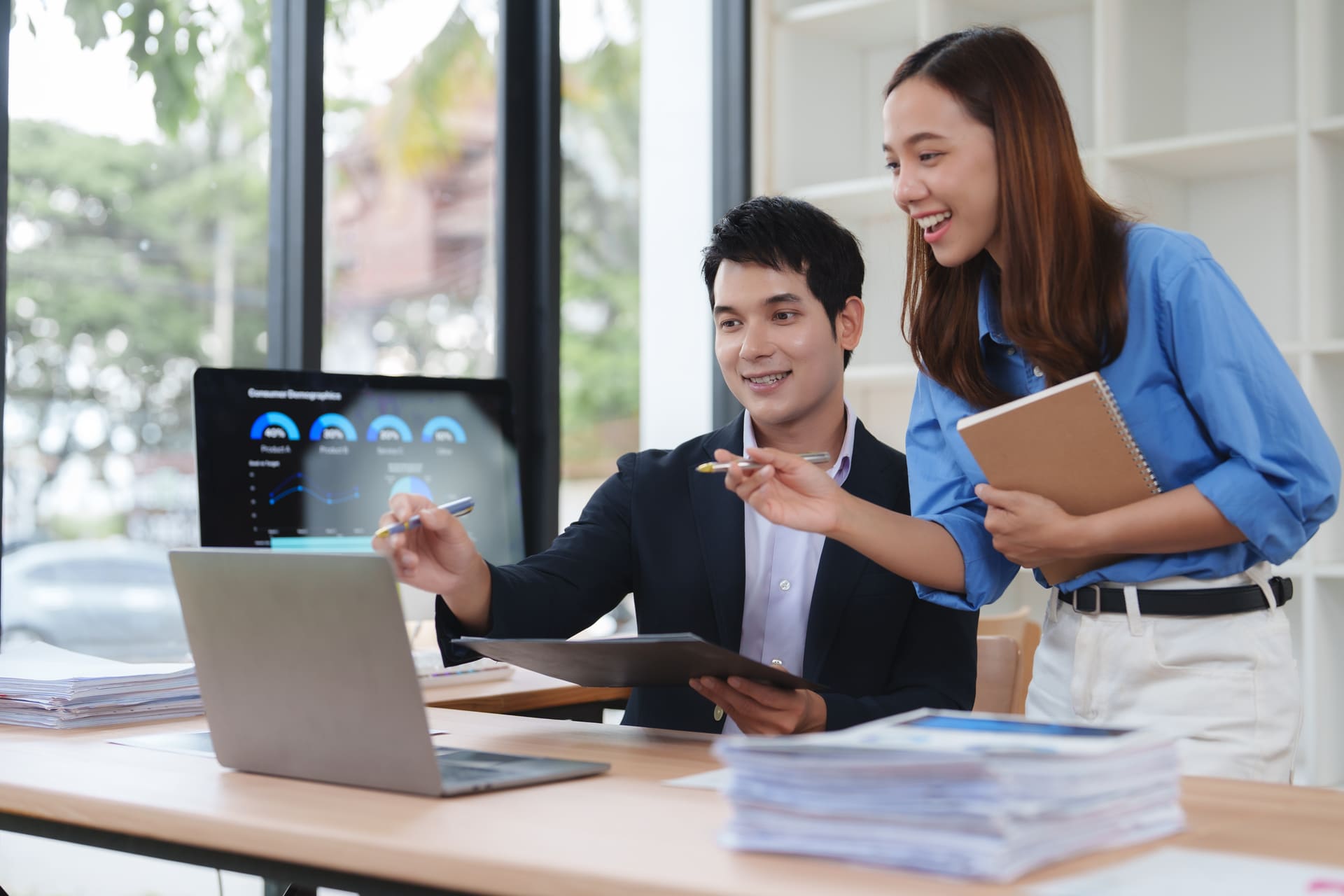Two coworkers analyze data together at a desk; the man points with a pen while the woman shares a notebook. The laptop displays graphs.