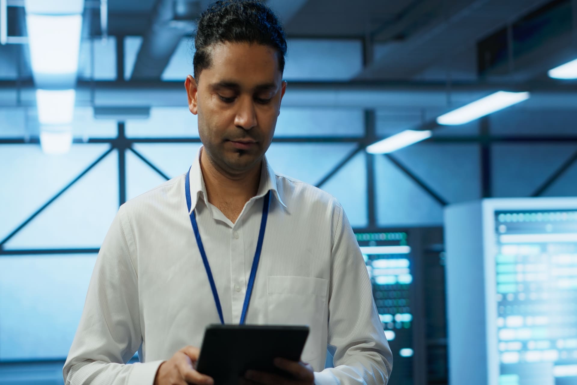 programmer-walking-in-high-tech-data-center-using tablet | CMIT Solutions Man in a white shirt with a blue lanyard stands in a data center, looking at a tablet in his hands.