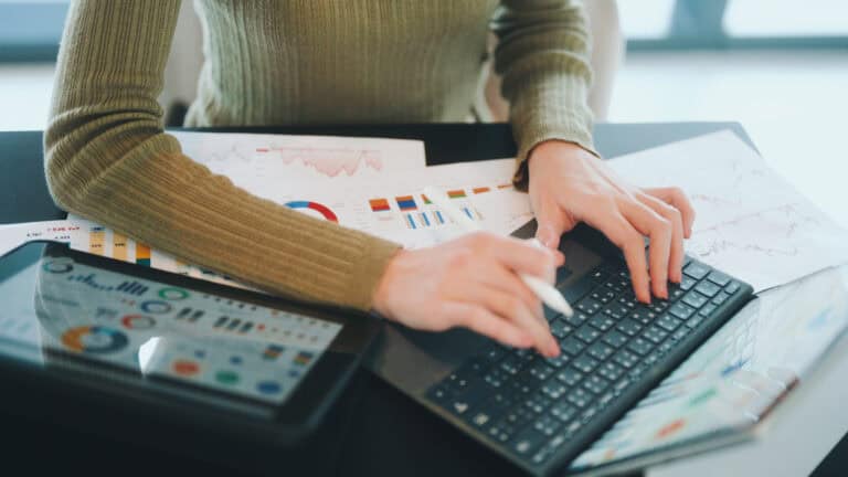 Person in a sweater typing on a laptop with financial charts and documents on the desk